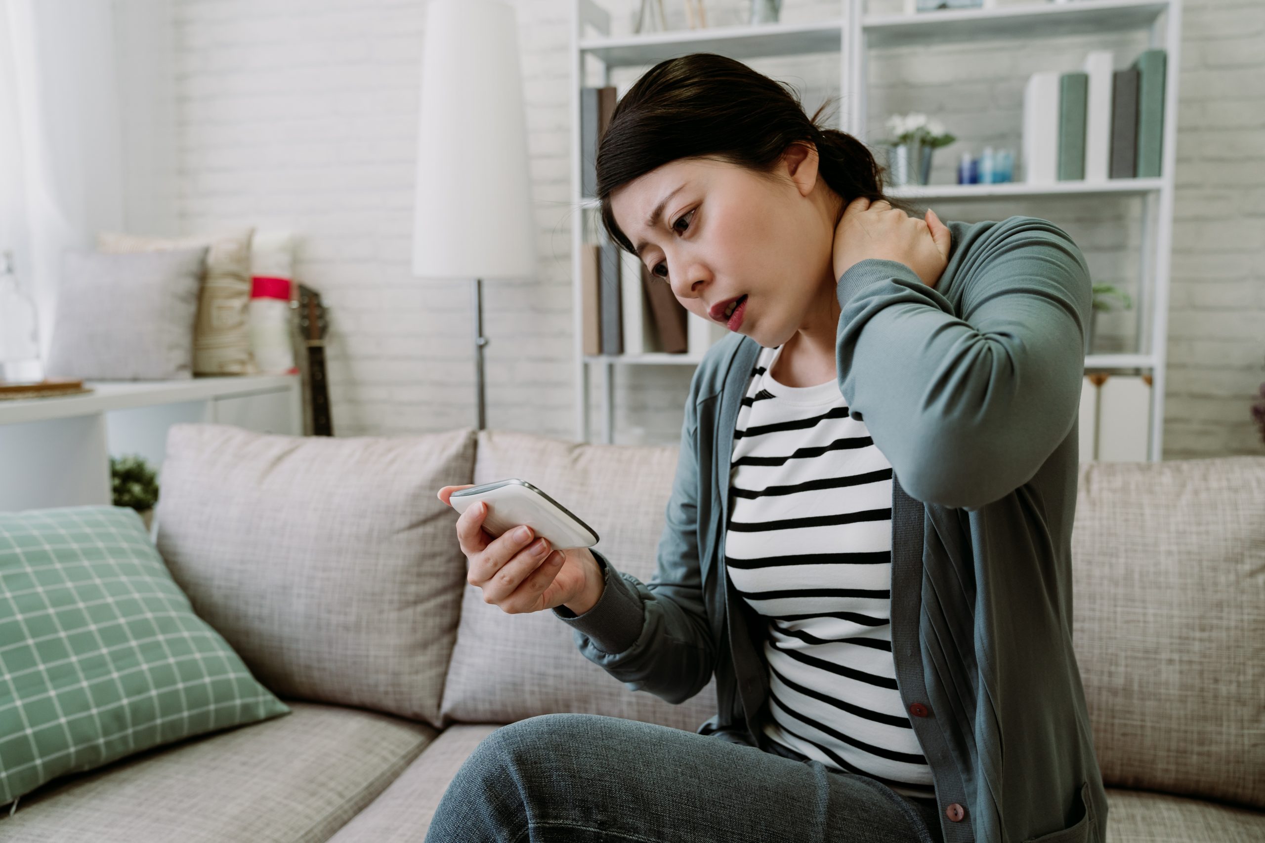 millennial woman tries to relive shoulder tension while looking at her phone screen