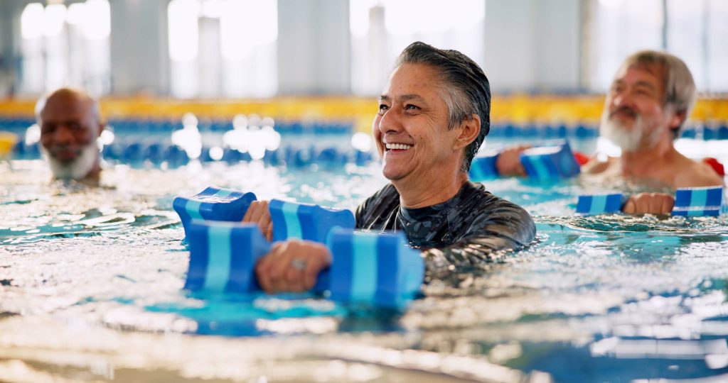 Older adults participating in a water aerobics class, using foam dumbbells for gentle, low-impact exercise in an indoor swimming pool.